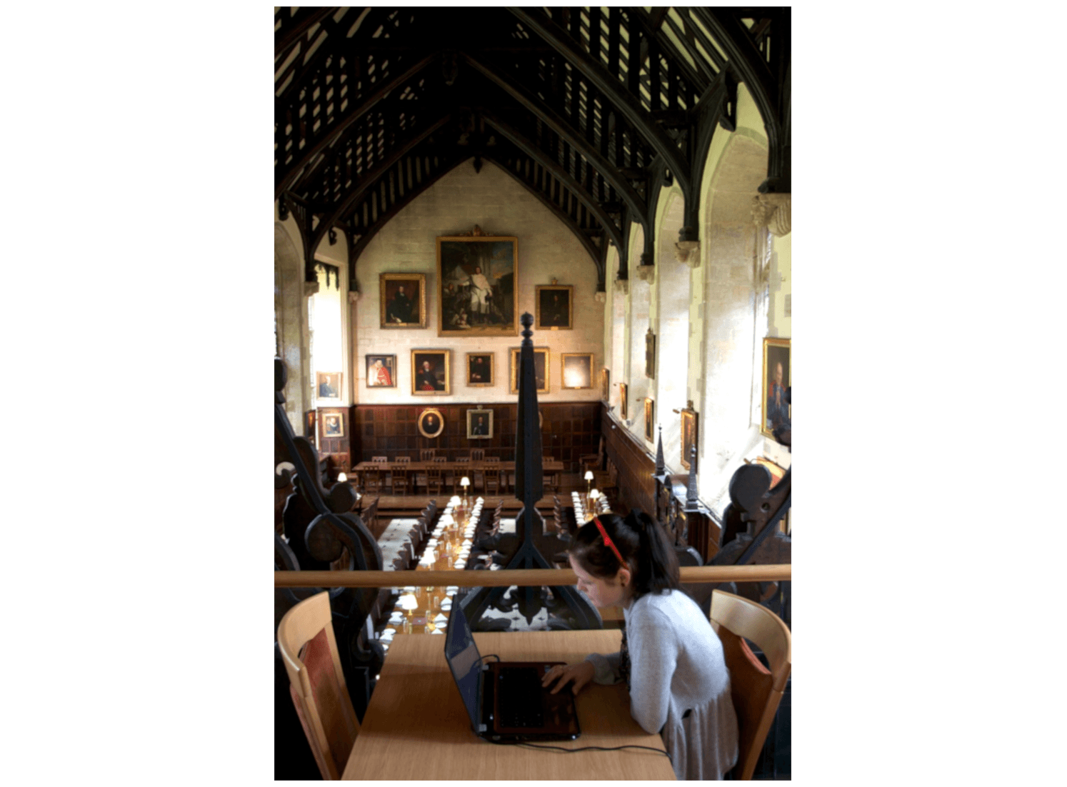 L'immagine mostra una persona seduta a un tavolo che lavora su un computer portatile, affacciata su una sala da pranzo o una sala conferenze in stile antico, adornata con ritratti alle pareti e un soffitto a volta in legno.