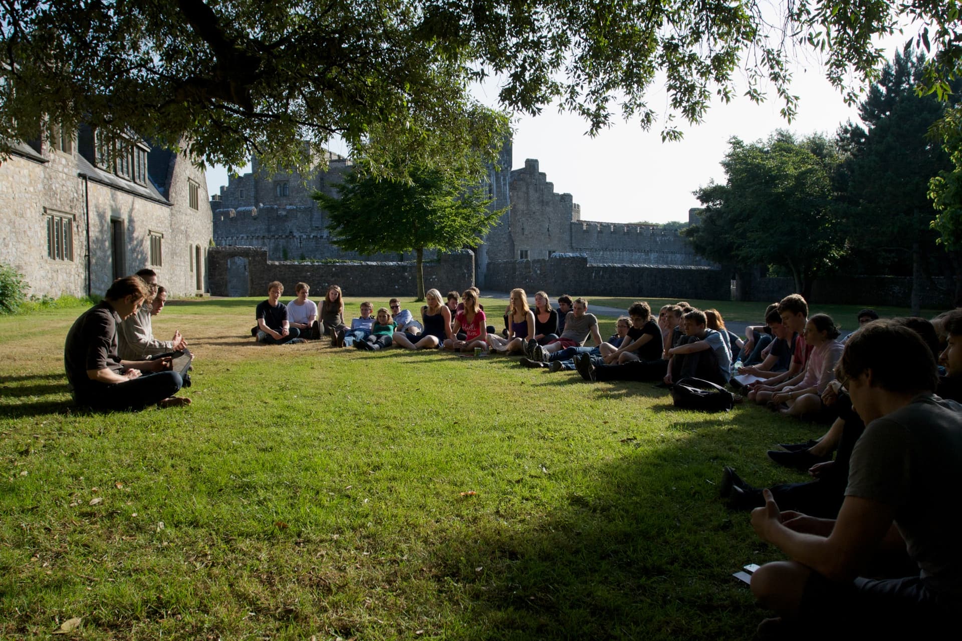 Un gruppo di persone è seduto in cerchio su un prato verde, ascoltando un oratore in un ambiente che sembra un campus storico o un parco con vecchi edifici in pietra sullo sfondo.