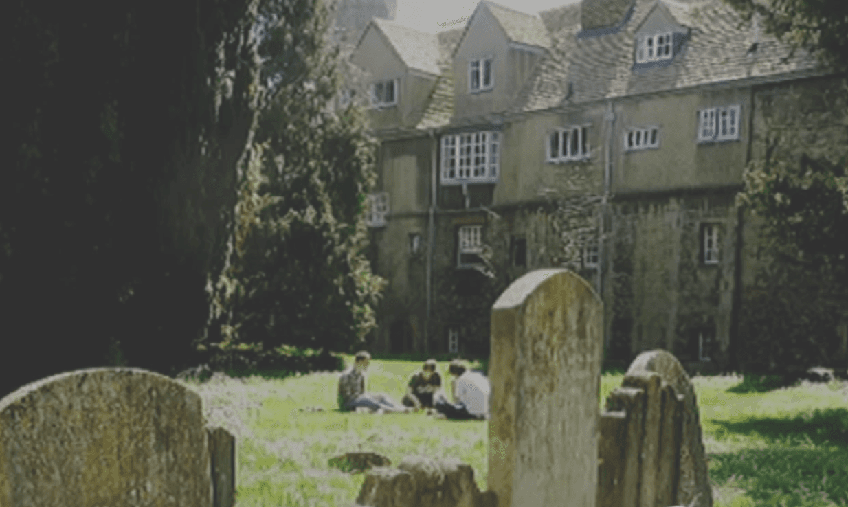 L'immagine mostra un gruppo di persone sedute sul prato davanti a un vecchio edificio, con lapidi di pietra in primo piano.