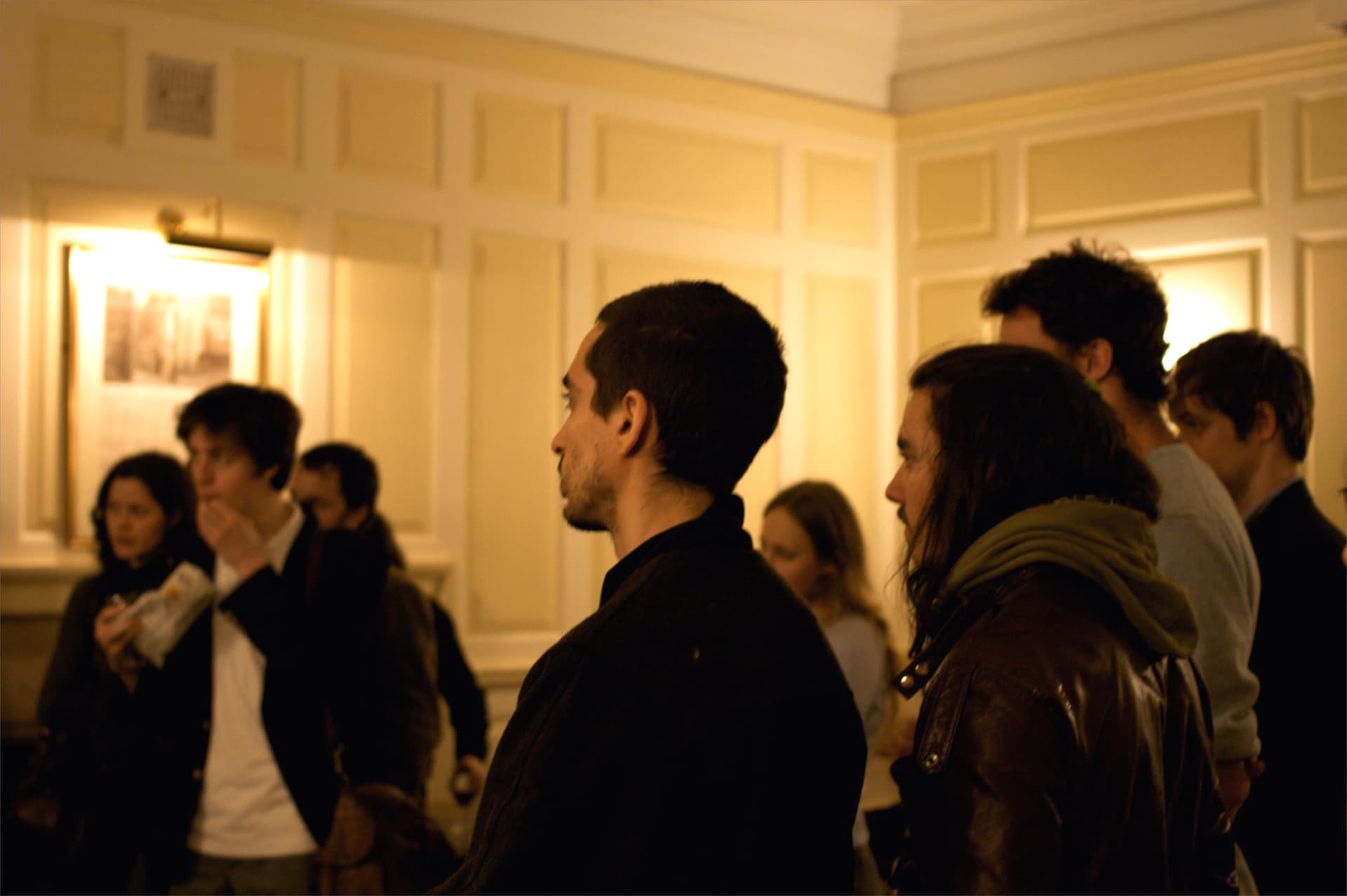 L'immagine mostra un gruppo di persone in uno spazio interno, forse durante un evento sociale o un incontro, con un giovane di profilo in primo piano che guarda verso destra.