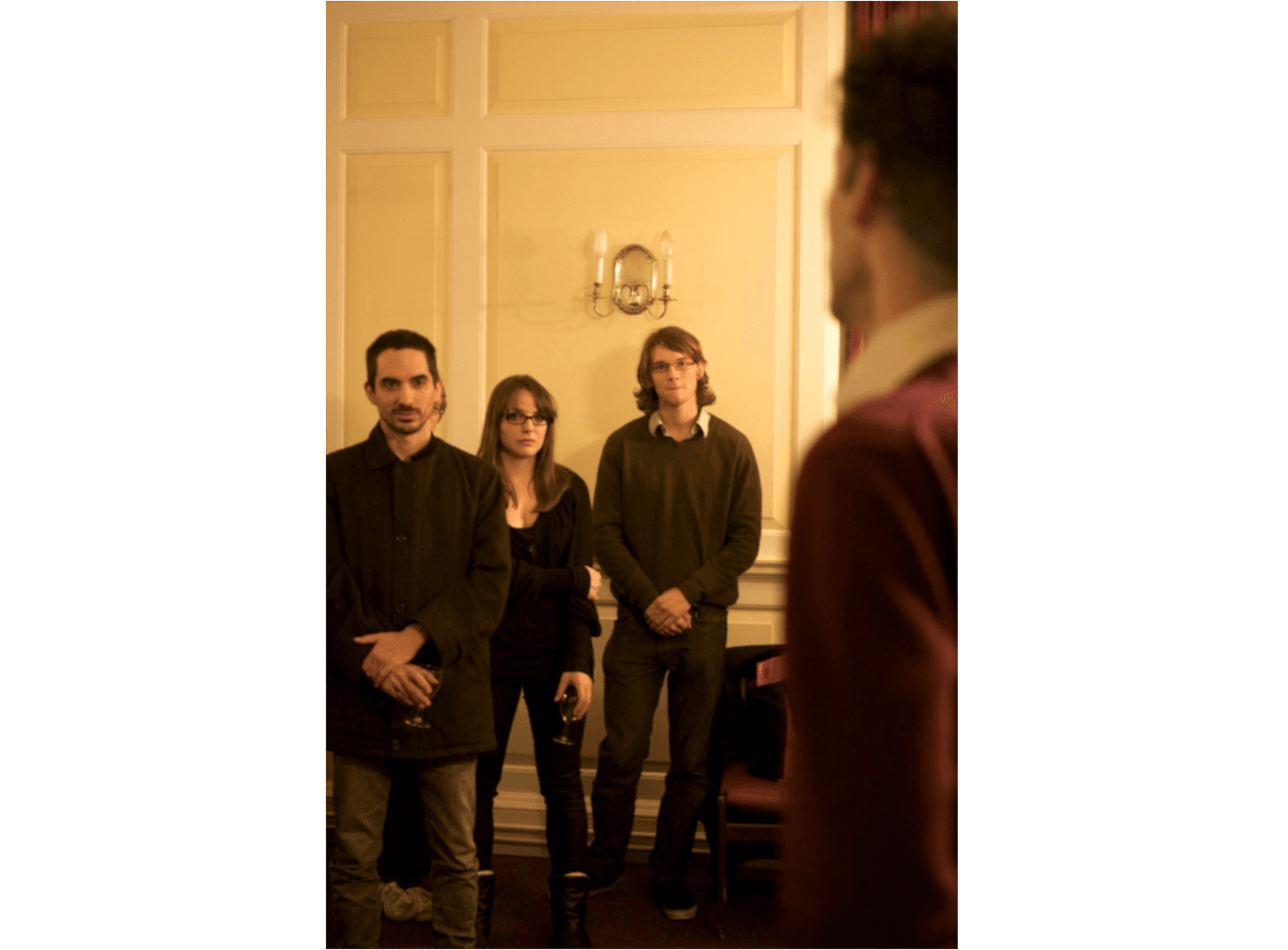 L'immagine mostra tre persone in piedi in una stanza arredata in stile classico, che guardano verso qualcosa o qualcuno fuori campo, in un'atmosfera rilassata.