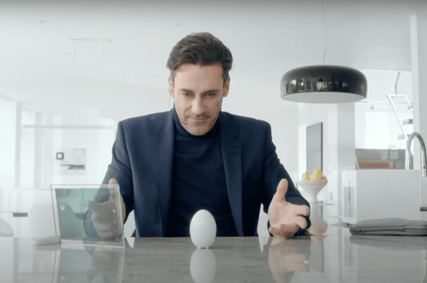 Un uomo in giacca e cravatta è chino su una superficie lucida in una cucina moderna e osserva con interesse un elettrodomestico a forma di uovo posizionato orizzontalmente. La scena suggerisce un'atmosfera di curiosità o sorpresa.