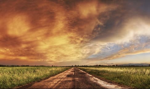 L'immagine mostra una strada diritta che attraversa una pianura, fiancheggiata da campi verdi sotto un cielo che appare drammatico sulla sinistra, con nuvole che riflettono toni arancioni e gialli, e limpido e luminoso sulla destra.