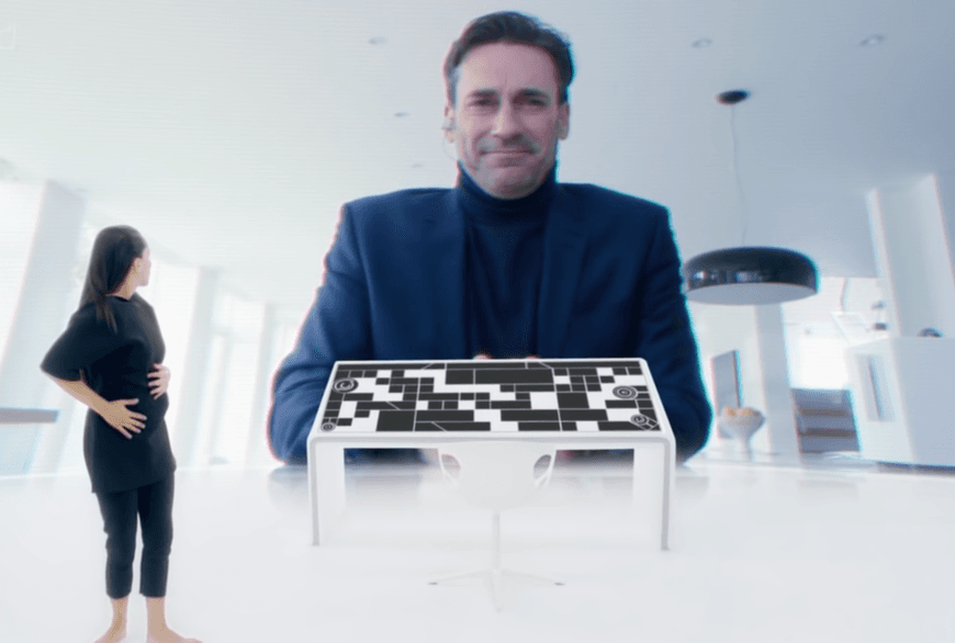 L'immagine mostra una donna in piedi davanti a un grande schermo, con un uomo sorridente che si protende verso di lei. Davanti alla donna c'è un tavolo di controllo in stile futuristico.