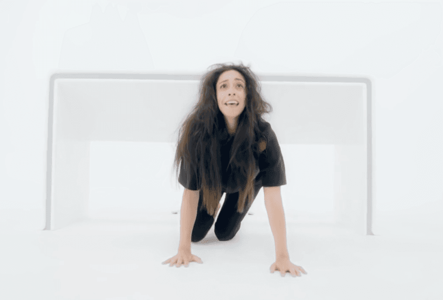 L'immagine mostra una donna inginocchiata con le mani appoggiate sul pavimento in una stanza bianca, con un'espressione che denota paura e sofferenza.